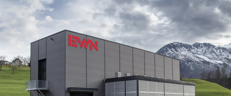 Grau verkleidetes längliches Gebäude mit Aufschrift EWN mit Wiesen und Bergen...