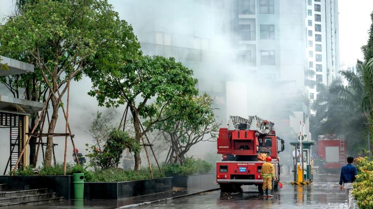 Feuerwehrauto mit Einsatzkraft vor einem rauchumgebenen Hochhaus mit Bäumen...