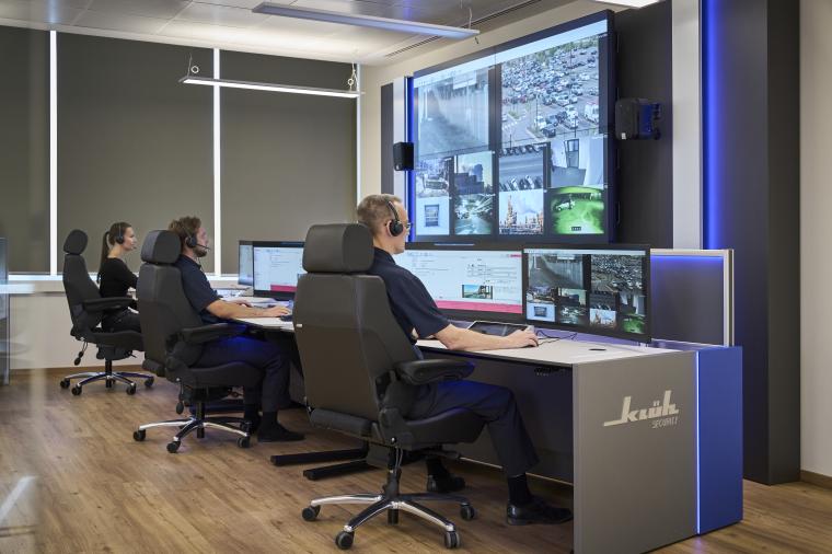 Drei Mitarbeiter arbeiten vor einer Reihe von Bildschirmen, dahinter ein...