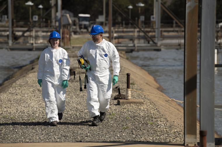 Two people in white protective clothing and helmets walk across an outdoor...