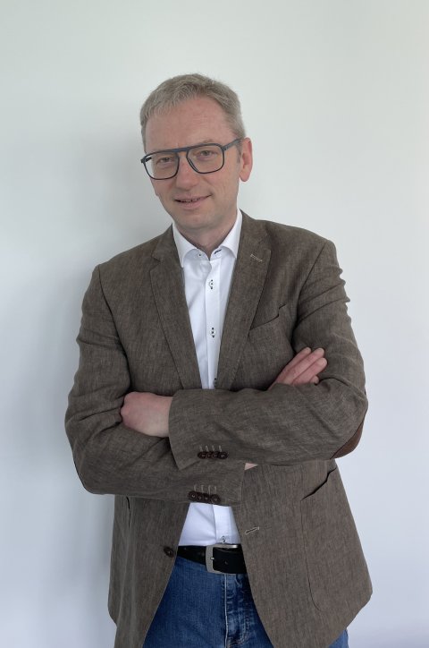 Person mit braunem Blazer, weißem Hemd und blauer Jeans vor weißem Hintergrund, mit verschränkten Armen; Gesicht unkenntlich