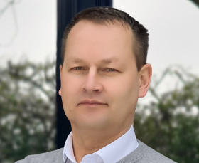 Person in grauem Pullover und weißem Hemd vor einem verschwommenen Hintergrund mit Bäumen