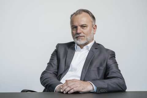 Person in grauem Sakko und weißem Hemd sitzt an einem Tisch vor hellem Hintergrund