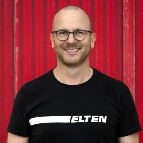 Person in schwarzem Elten T Shirt vor rotem metallischen Hintergrund