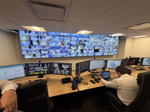 A man sitting in front of several monitors, with a wall of split screens behind him